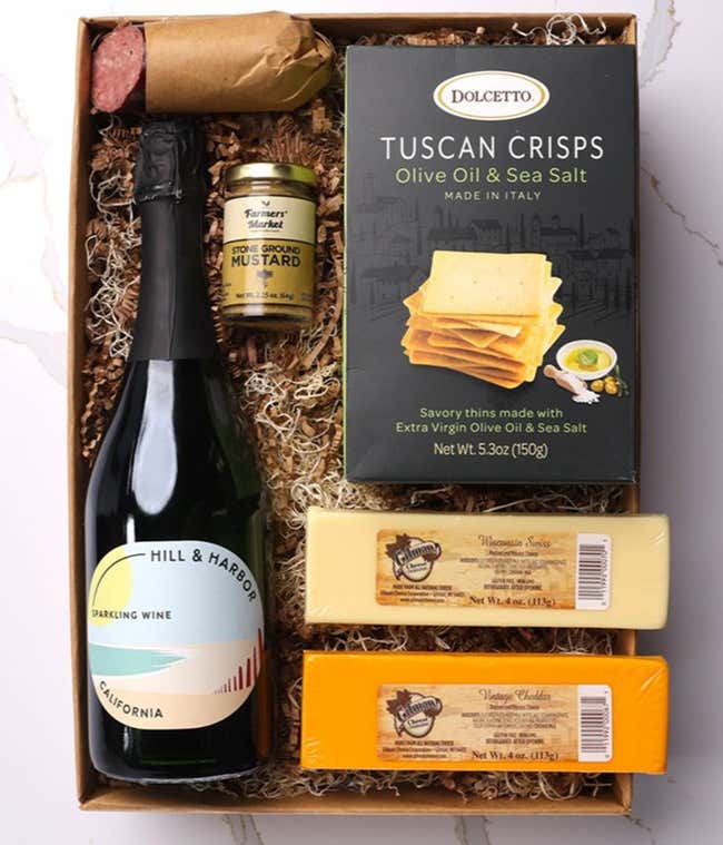 A gift box with Tuscan crisps, salami, cheeses, mustard, and a bottle of Hill &amp; Harbor sparkling wine. Surrounding the box are plates with cheese, salami, mustard, and two glasses of sparkling wine.