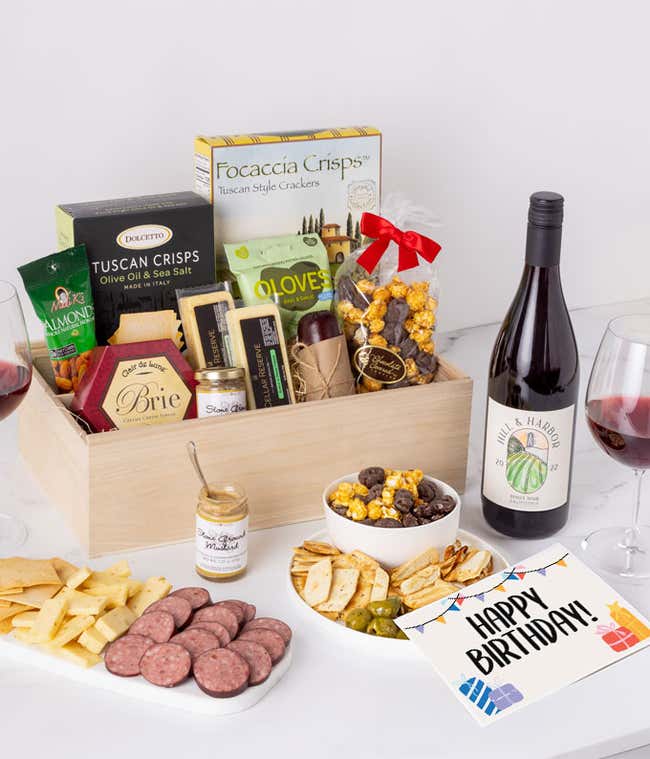 A charcuterie gift set with Tuscan crisps, focaccia crackers, 3 cheeses, summer sausage, olives, mustard, smoked almonds, chocolate-covered popcorn, and a birthday card.
