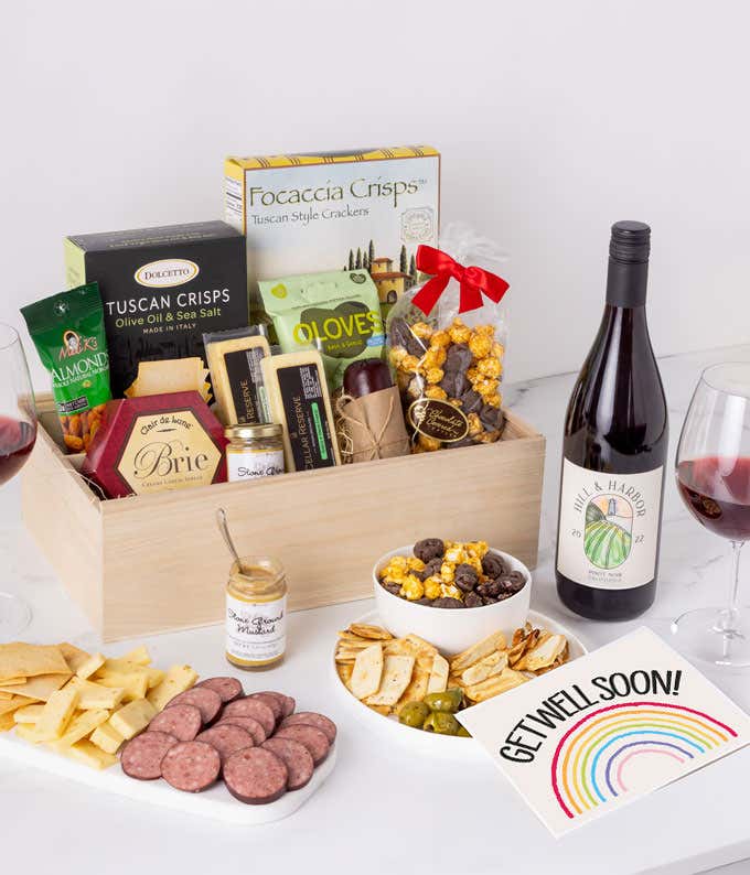 A charcuterie gift set with Tuscan crisps, focaccia crackers, 3 cheeses, summer sausage, olives, mustard, smoked almonds, chocolate-covered popcorn, a bottle of Pinot Noir, and a get well soon card.