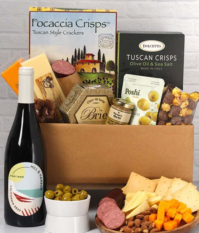 A charcuterie gift set with Tuscan crisps, focaccia crackers, 3 cheeses, olives, mustard, sausage almonds, chocolate-covered popcorn and a bottle of Pinot Noir.