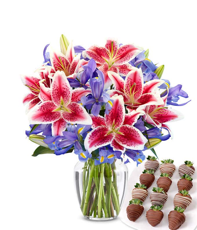 Clear glass vase filled with a vibrant bouquet of hot pink stargazer lilies and deep violet-blue irises, paired with a plate of swizzled chocolate-covered strawberries drizzled in dark and white chocolate.