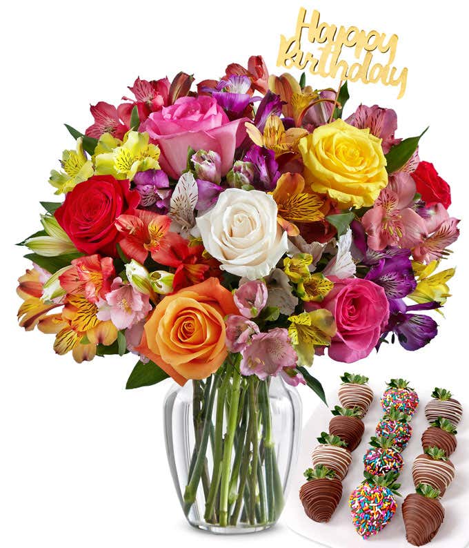 Flower arrangement with mixed roses and multicolor alstroemeria in a clear glass vase, accented with a gold Happy Birthday pick and paired with chocolate-covered strawberries in festive drizzle and rainbow sprinkle designs.