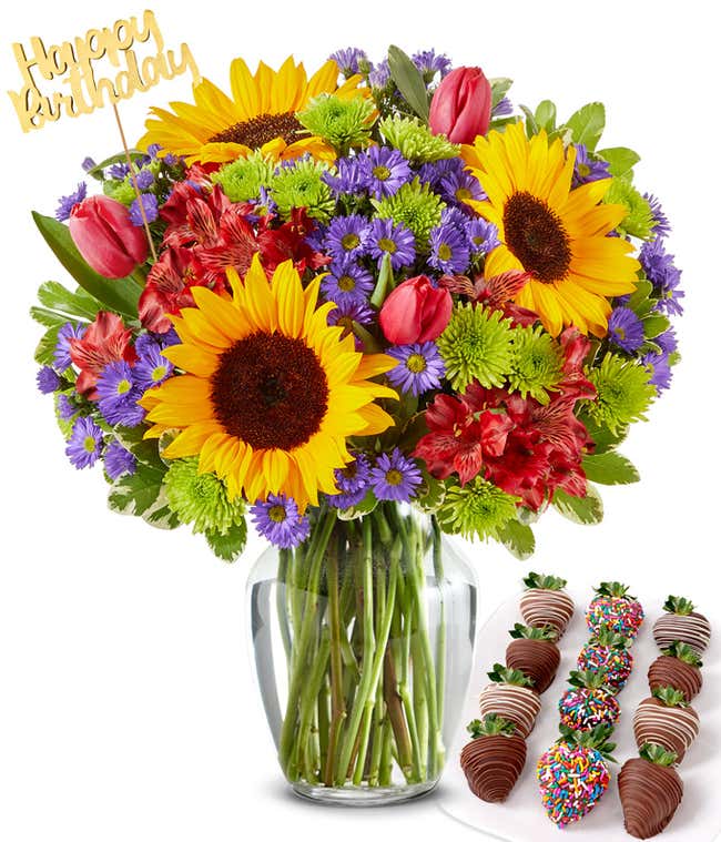 Flower arrangement with sunflowers, red alstroemeria, green poms, purple asters, and red tulips in a clear glass vase, with a gold Happy Birthday pick and paired with chocolate-covered strawberries, including rainbow sprinkle and drizzles.