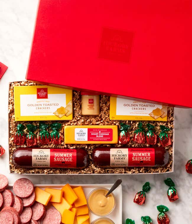 Red gift box filled with savory snacks and sweets including summer sausage, golden toasted crackers, smoked cheddar blend, sweet hot mustard, strawberry bon bons, displayed with sliced sausage, cheese, crackers, and mustard on a marble surface.