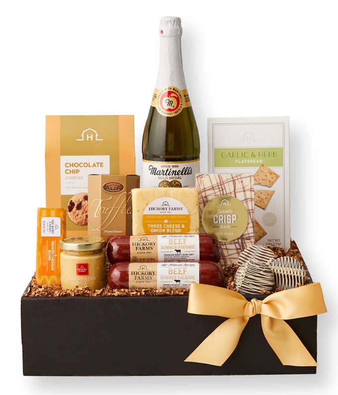 Gift basket with sparkling cider, chocolate chip cookies, crackers, cheese spreads, sausage, garlic & herb flatbread, and truffles in a black box with a gold bow, surrounded by a serving of sausage, cheese, and crackers on a marble surface.