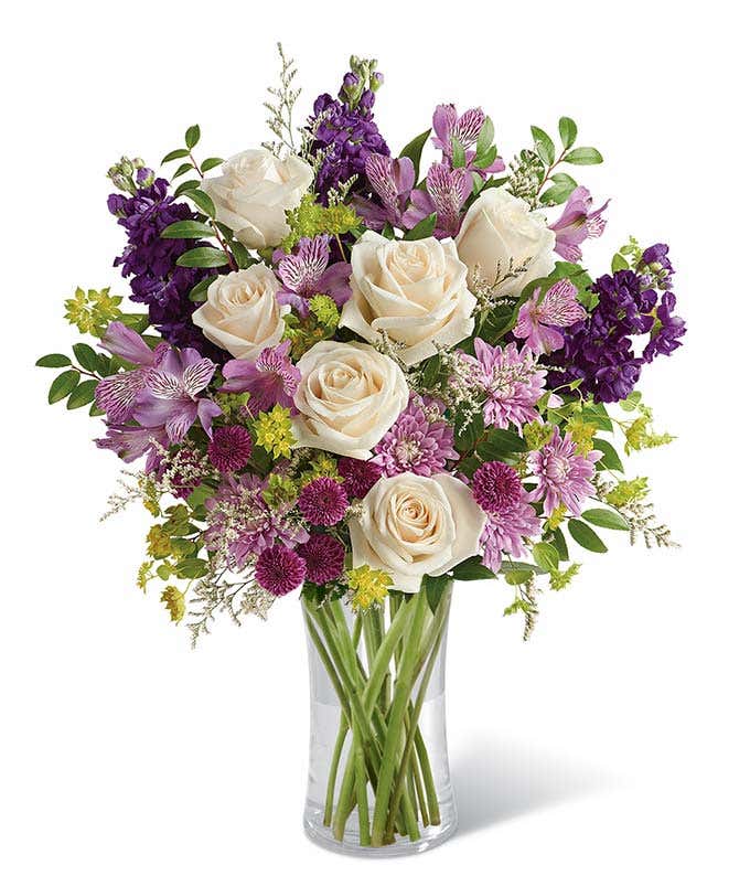 A sophisticated floral arrangement featuring crème roses, lavender alstroemeria, purple stock, purple button chrysanthemums, lavender cushion spray chrysanthemums, white limonium, bupleurum, and huckleberry, all elegantly arranged in a cylinder vase.