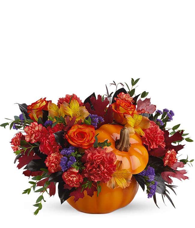 A vibrant floral arrangement featuring orange carnations, orange mini carnations, orange Peruvian lilies, purple statice, deluxe and premium bi-color roses, and oak leaves, all beautifully presented in a keepsake container.