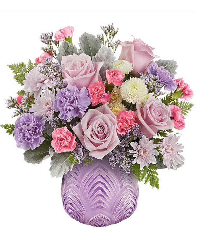 Soft pastel floral arrangement featuring lavender roses, lavender carnations, pink miniature carnations, lavender cushion spray chrysanthemums, white button spray chrysanthemums, dusty miller, and lush greenery in a textured lavender glass vase.