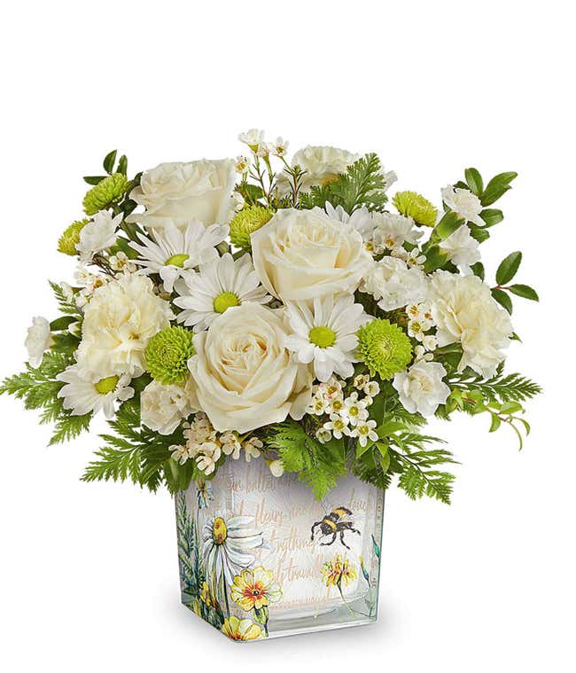 White roses, white daisies, white carnations, green button mums, white waxflower, and assorted greenery arranged in a square glass container with a watercolor-style bee and floral design.
