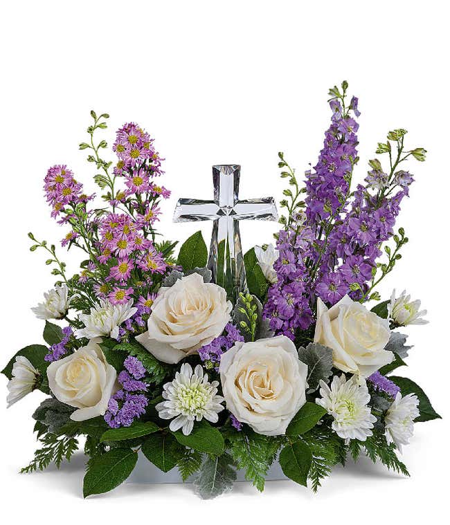 A graceful floral arrangement showcasing cr&egrave;me roses, lavender larkspur, lavender asters, lavender statice, white mums, and floral greens, elegantly accented with a crystal cross. This bouquet blends soft, calming colors for a serene display.