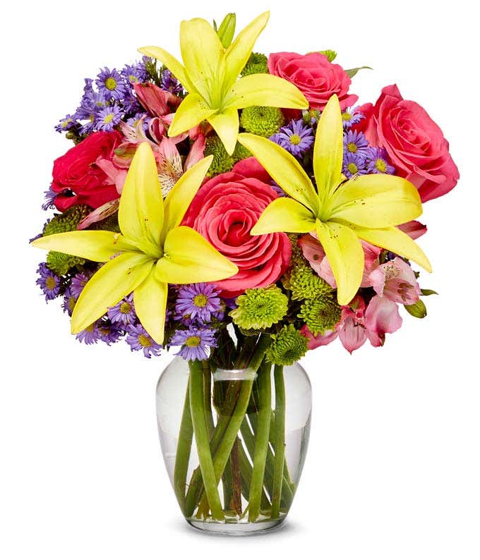 A bouquet featuring yellow lilies, pink roses, green poms, and purple monte cassino, arranged in a clear fluted vase.