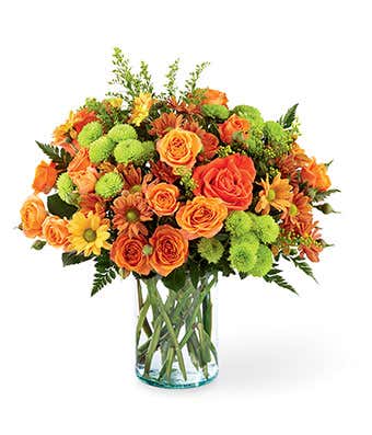 A vibrant floral arrangement featuring orange roses, orange spray roses, butterscotch daisies, green button poms, and solidago, all beautifully displayed in a clear glass vase.