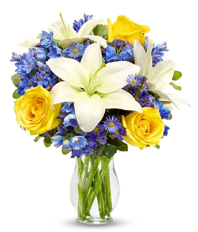 Yellow roses, blue delphinium and white lilies in a clear glass vase