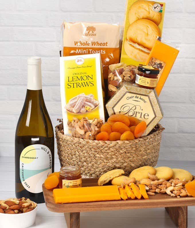 A two-toned woven basket filled with two cheeses, lemon flavored baked goods, crackers, dried apricots, gourmet nuts, and honey, paired with a bottle of red wine.