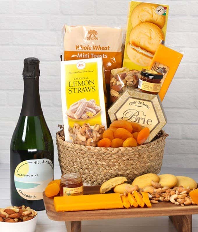 A two-toned woven basket filled with two cheeses, lemon flavored baked goods, crackers, dried apricots, gourmet nuts, and honey, paired with a bottle of sparkling wine.
