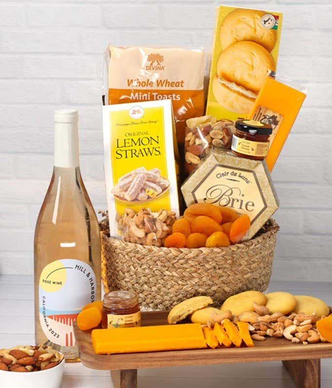 A two-toned woven basket filled with two cheeses, lemon flavored baked goods, crackers, dried apricots, gourmet nuts, and honey, paired with a bottle of rosé wine.