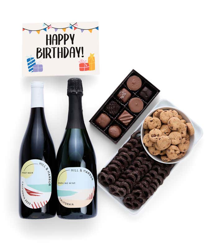 A birthday gift set with two bottles of Hill & Harbor wine (Pinot Noir and Sparkling Wine), a box of assorted chocolates, a tray of chocolate-covered pretzels, a bowl of mini chocolate chip cookies, and a birthday message card.