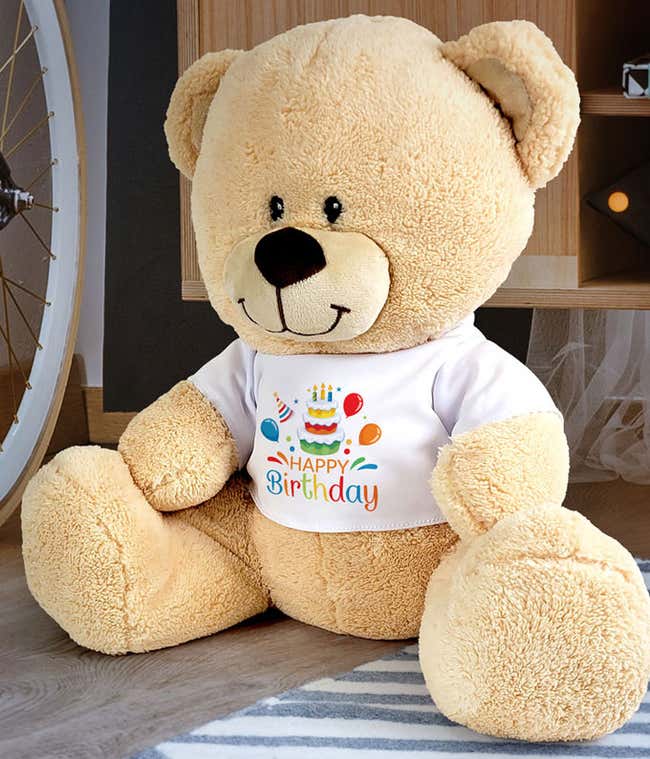 Plush tan teddy bear wearing a white t-shirt printed with a colorful birthday cake, balloons, and Happy Birthday text, shown seated against a white background.
